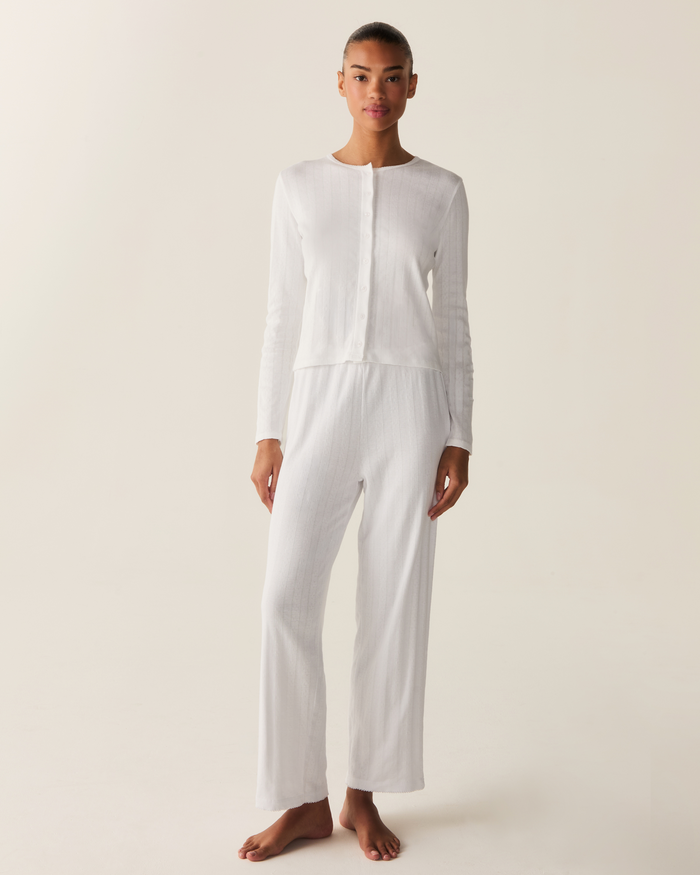 A person stands against a plain background, wearing the Women’s Pointelle Cardigan in White paired with loose white pants. They are barefoot and facing forward with a neutral expression.