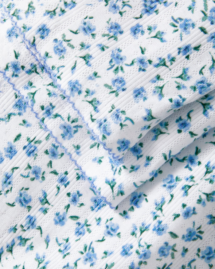 A woman stands barefoot against a plain background, wearing the Women's Pointelle Pant in Blue Floral paired with a matching short-sleeve pajama top featuring a white base and small blue floral print.