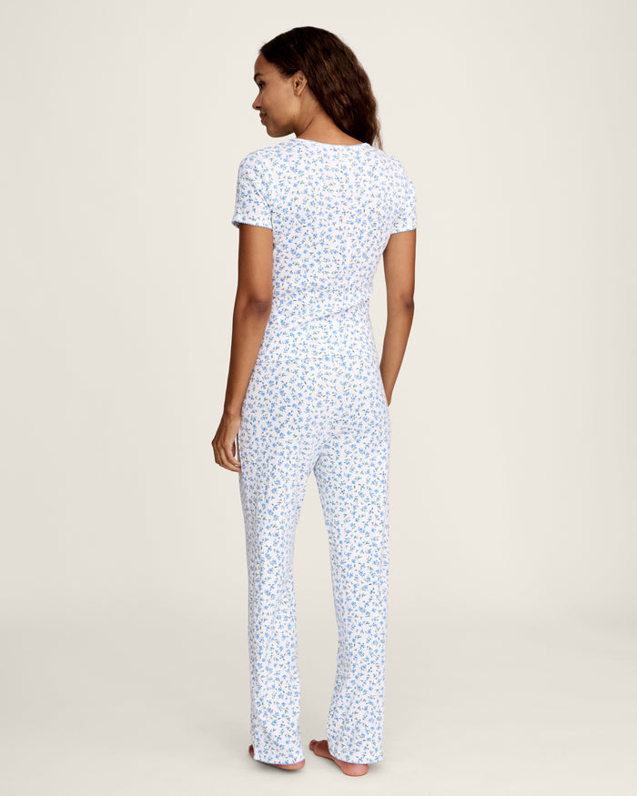 A woman stands barefoot against a plain background, wearing the Women's Pointelle Pant in Blue Floral paired with a matching short-sleeve pajama top featuring a white base and small blue floral print.
