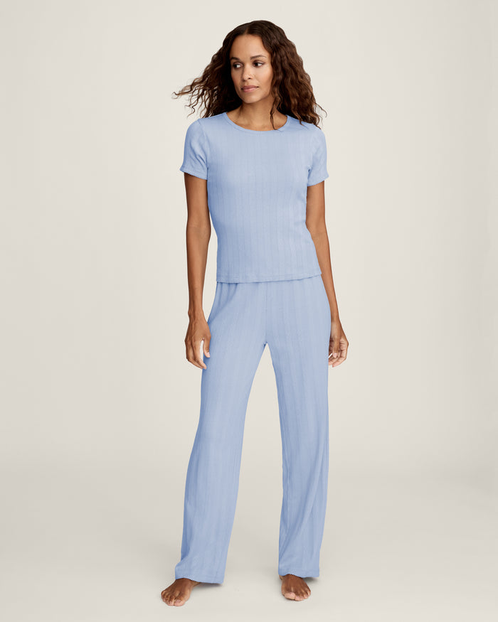 A woman stands barefoot in Women's Pointelle Pant in Periwinkle, paired with a matching short-sleeve top. She has wavy brown hair and gazes to the side against a plain, light background.