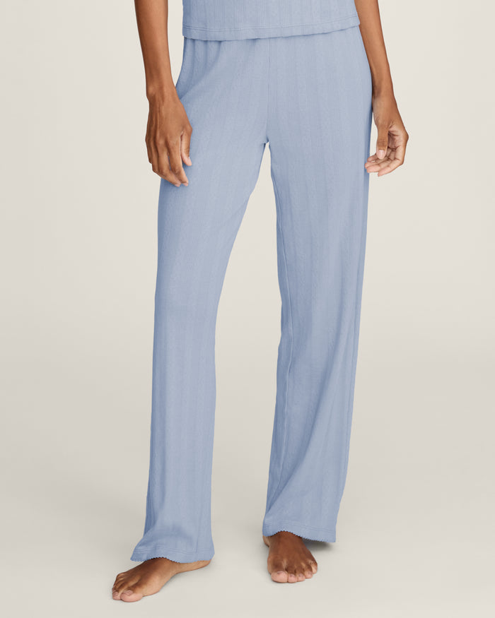 A woman stands barefoot in Women's Pointelle Pant in Periwinkle, paired with a matching short-sleeve top. She has wavy brown hair and gazes to the side against a plain, light background.