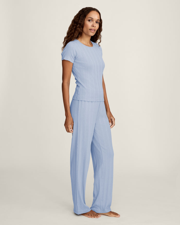 A woman stands barefoot in Women's Pointelle Pant in Periwinkle, paired with a matching short-sleeve top. She has wavy brown hair and gazes to the side against a plain, light background.