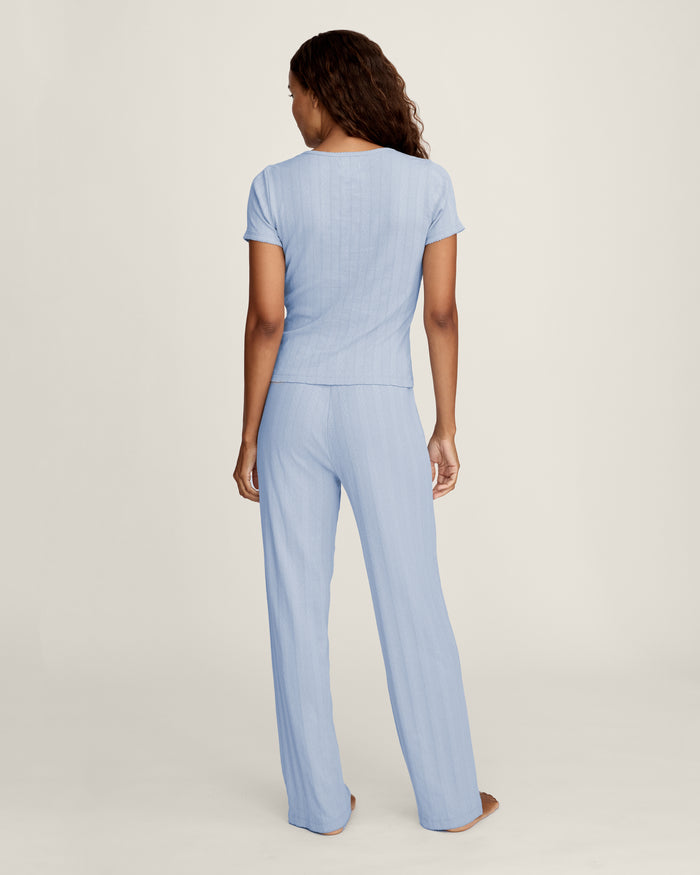 A woman stands barefoot in Women's Pointelle Pant in Periwinkle, paired with a matching short-sleeve top. She has wavy brown hair and gazes to the side against a plain, light background.