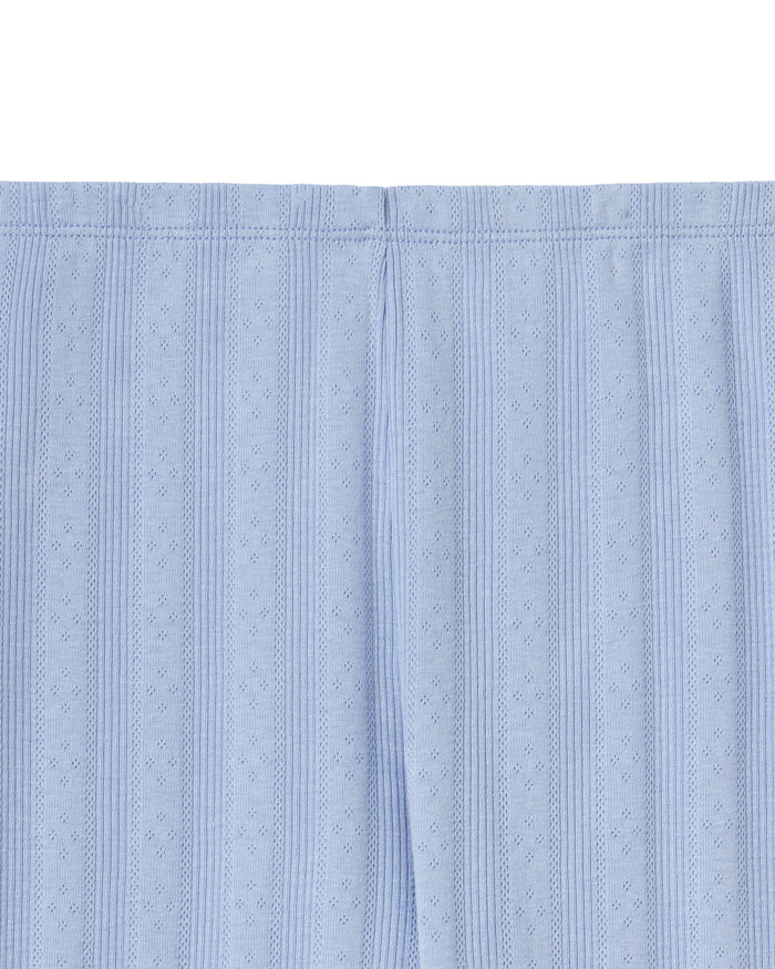 A woman stands barefoot in Women's Pointelle Pant in Periwinkle, paired with a matching short-sleeve top. She has wavy brown hair and gazes to the side against a plain, light background.