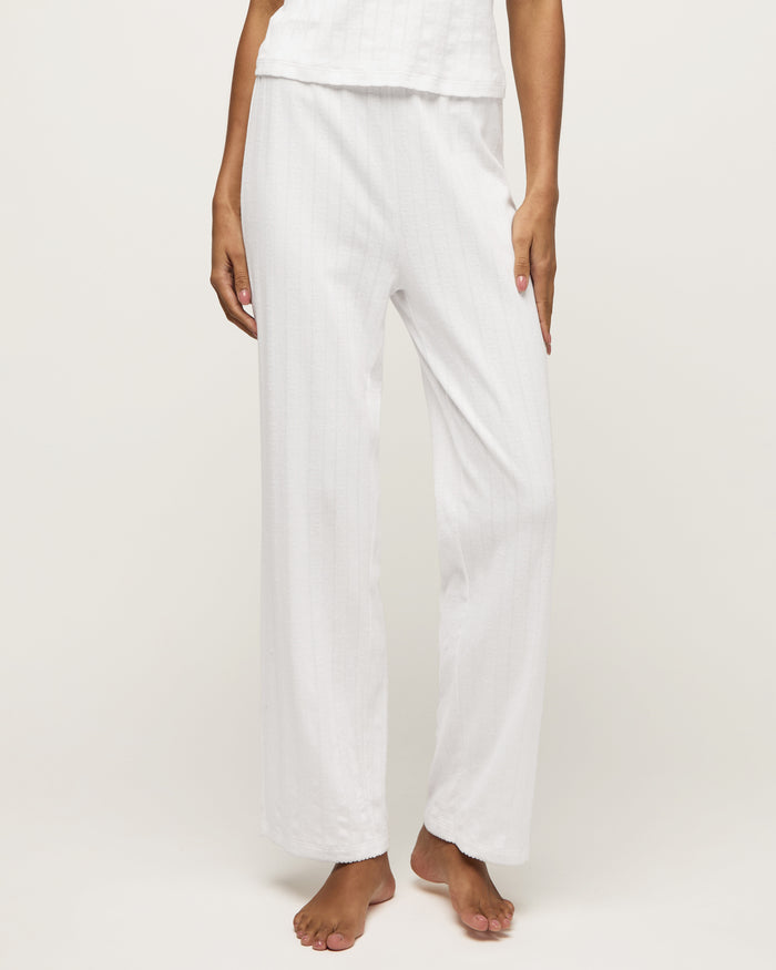 A woman stands barefoot on a light background wearing the Women's Pointelle Pant in White, paired with a sleeveless white tank top, posing with a relaxed and neutral expression.