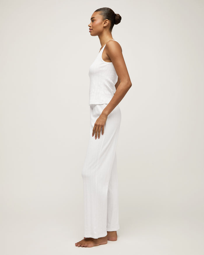 A woman stands barefoot on a light background wearing the Women's Pointelle Pant in White, paired with a sleeveless white tank top, posing with a relaxed and neutral expression.