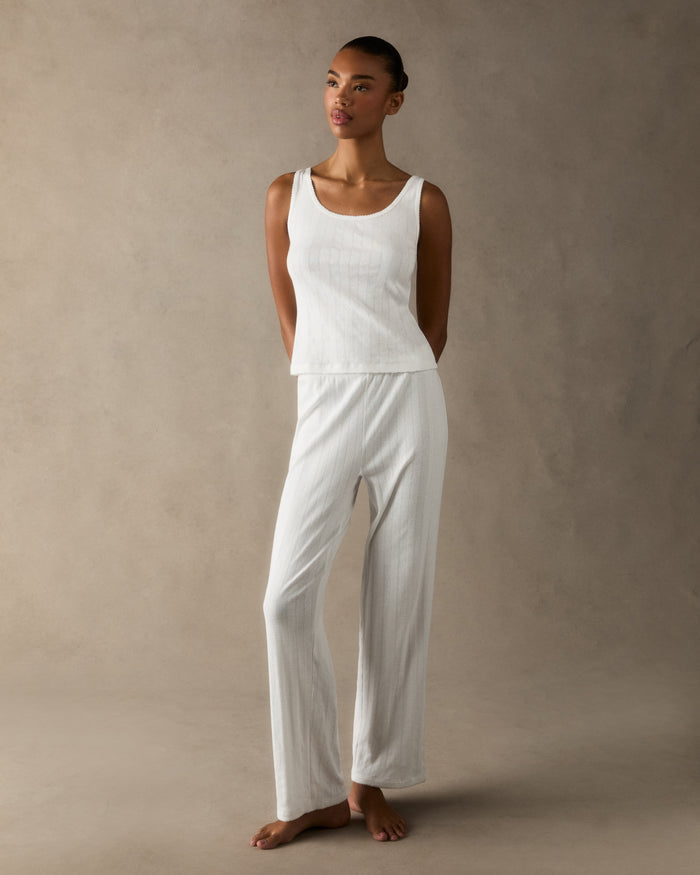A woman stands barefoot on a light background wearing the Women's Pointelle Pant in White, paired with a sleeveless white tank top, posing with a relaxed and neutral expression.