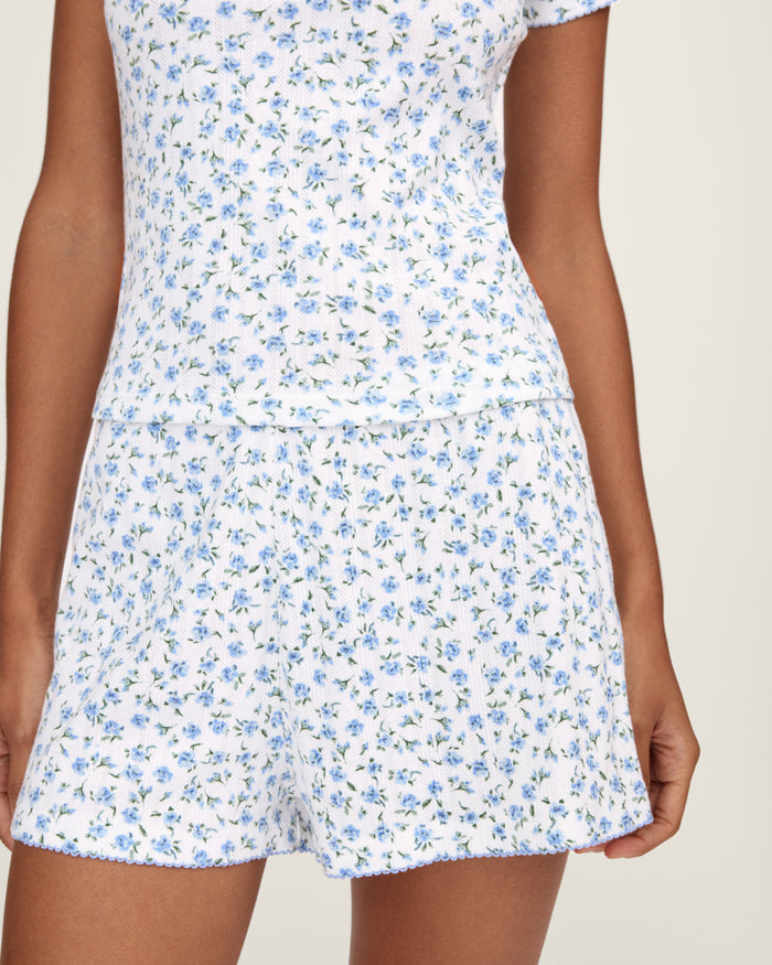 A person stands barefoot against a plain background, wearing Women's Pointelle Shorts in Blue Floral with a matching short-sleeve top. They have long, wavy hair and a relaxed, neutral expression.