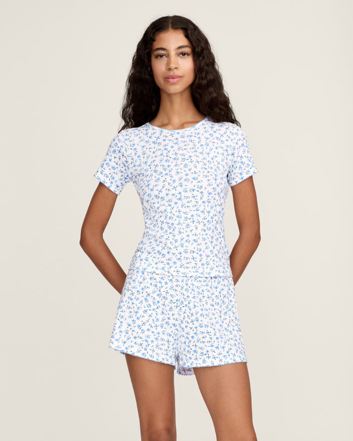 A person stands barefoot against a plain background, wearing Women's Pointelle Shorts in Blue Floral with a matching short-sleeve top. They have long, wavy hair and a relaxed, neutral expression.