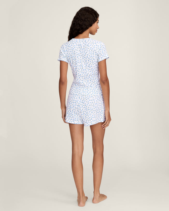 A person stands barefoot against a plain background, wearing Women's Pointelle Shorts in Blue Floral with a matching short-sleeve top. They have long, wavy hair and a relaxed, neutral expression.