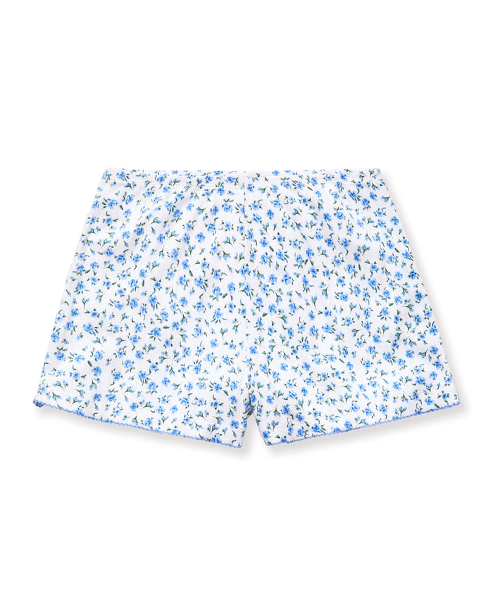 A person stands barefoot against a plain background, wearing Women's Pointelle Shorts in Blue Floral with a matching short-sleeve top. They have long, wavy hair and a relaxed, neutral expression.