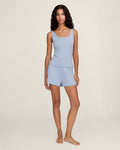 A woman with long wavy hair stands barefoot on a plain background, wearing Women's Pointelle Shorts in Periwinkle and a matching light blue ribbed tank top. She has a relaxed, neutral expression.