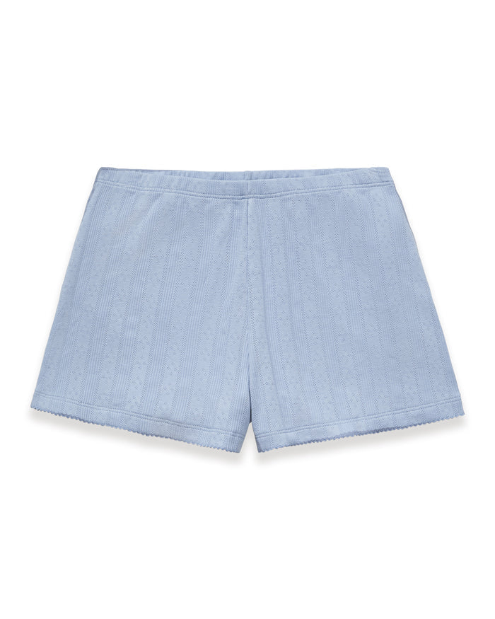A woman with long wavy hair stands barefoot on a plain background, wearing Women's Pointelle Shorts in Periwinkle and a matching light blue ribbed tank top. She has a relaxed, neutral expression.