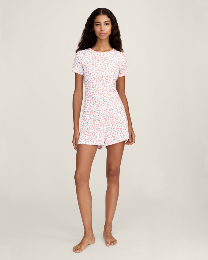 A woman with long curly hair and a relaxed expression stands barefoot against a neutral background, wearing Women's Pointelle Shorts in Pink Floral as part of a matching short-sleeved pajama set.