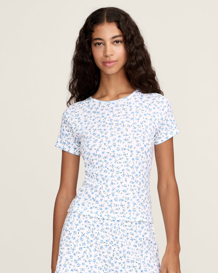 A young woman with long curly brown hair wears the Women's Pointelle T-Shirt in Blue Floral, paired with a matching skirt, against a plain light background.