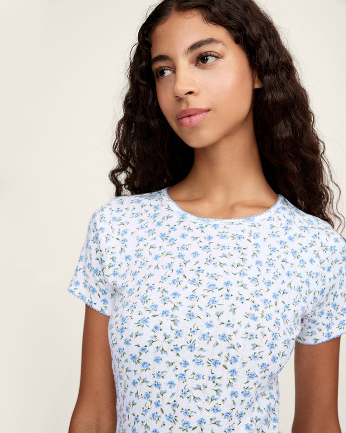 A young woman with long curly brown hair wears the Women's Pointelle T-Shirt in Blue Floral, paired with a matching skirt, against a plain light background.