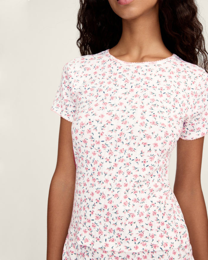 A woman with long curly hair wears the Women's Pointelle T-Shirt in Pink Floral, standing against a plain light background.