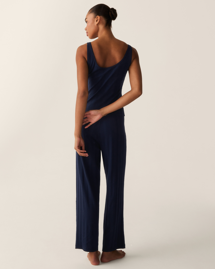 A woman stands barefoot, facing away, wearing the Women’s Pointelle Tank in Navy. Her hair is styled in a neat bun, and she rests one hand on her lower back against a plain, light background.