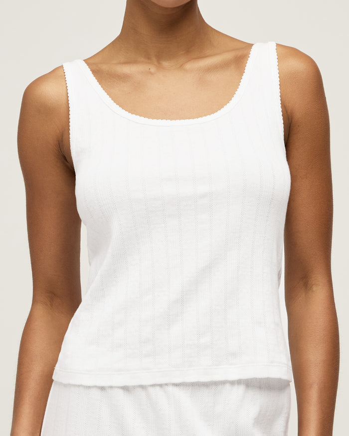 A woman with pulled-back hair wears the Women's Pointelle Tank in White and matching bottoms, standing against a plain light background and smiling softly at the camera.