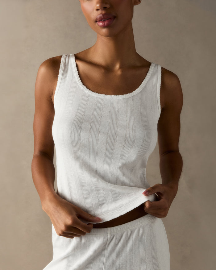 A woman with pulled-back hair wears the Women's Pointelle Tank in White and matching bottoms, standing against a plain light background and smiling softly at the camera.