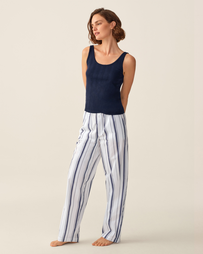 A woman stands barefoot against a plain light background, arms behind her back, wearing a navy sleeveless top and the Women’s Poplin Romy Pants in Parlor Stripe—white pajama pants with vertical blue stripes.