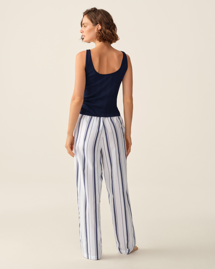 A person stands facing away, wearing a navy sleeveless top and the Women’s Poplin Romy Pants in Parlor Stripe — loose white pants with vertical blue stripes — against a light beige background.
