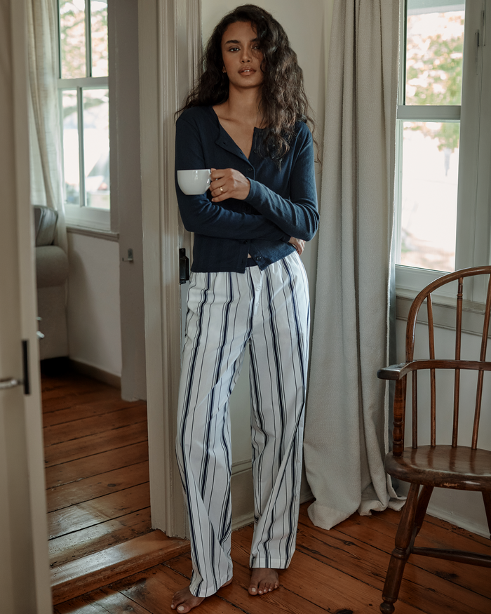 A woman with long curly hair stands barefoot indoors, holding a white mug. She wears a navy top and the Women’s Poplin Romy Pants in Parlor Stripe as sunlight streams through the windows behind her.