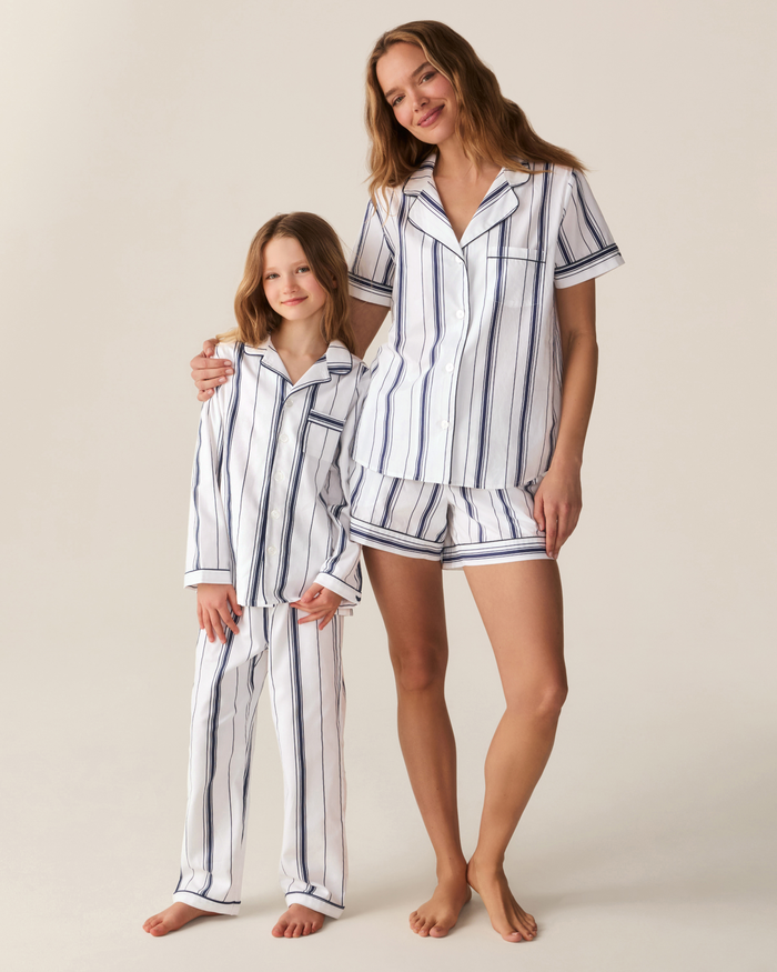 A woman and a child stand smiling, both dressed in matching white pajamas with navy parlor stripes. The woman wears the Women’s Poplin Short Sleeve Short Set in Parlor Stripe, while the child wears long pants.