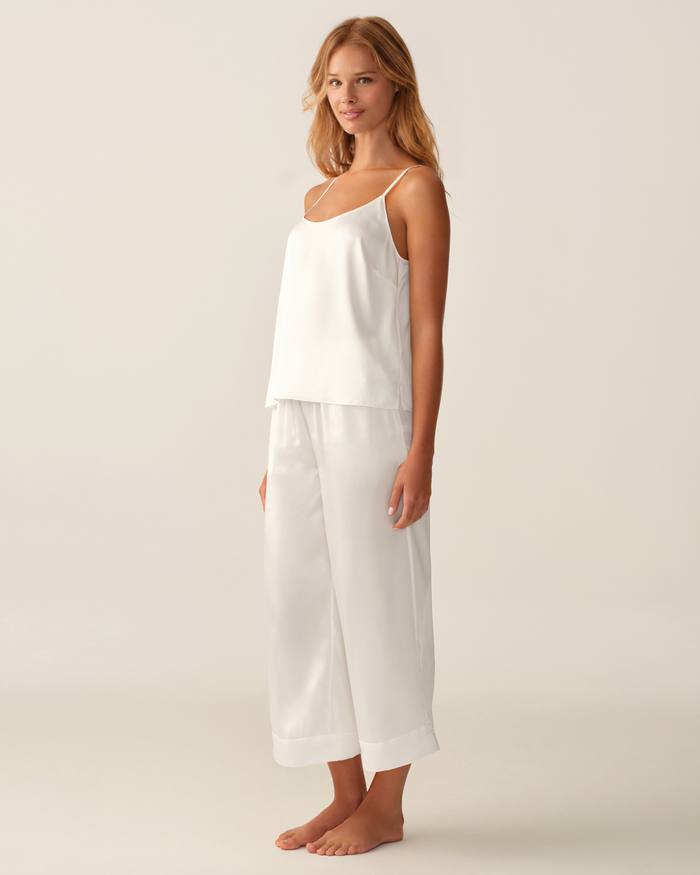A person stands barefoot on a light background, wearing the Women’s Silk Cami in White paired with matching wide-leg pants. They are turned slightly to the side and smiling gently.