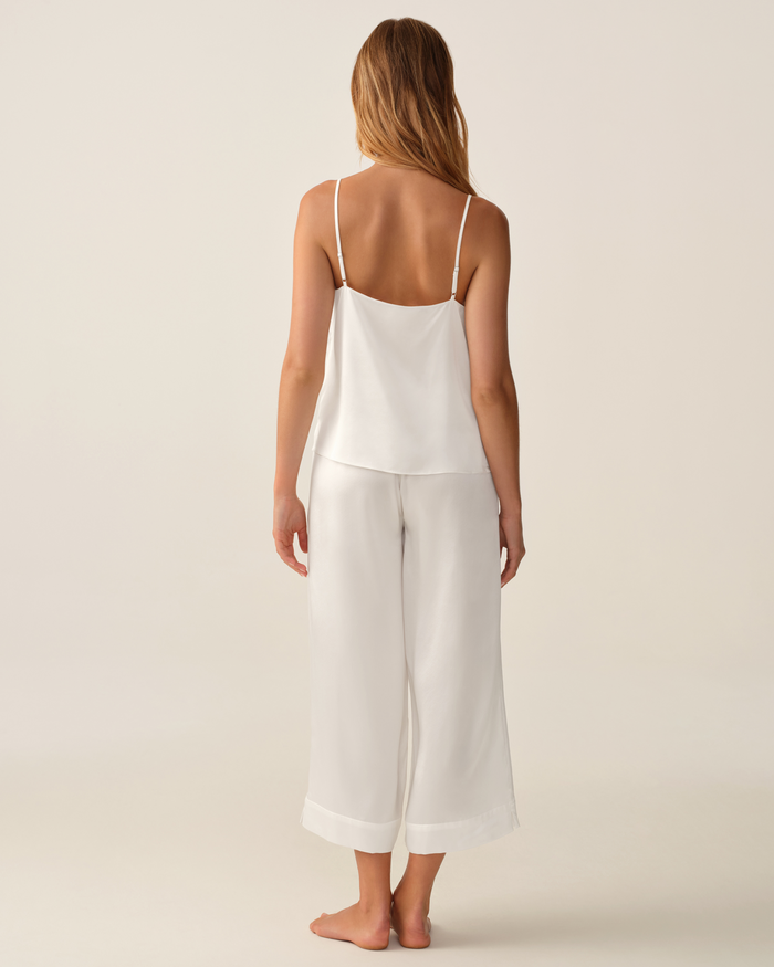 A woman stands barefoot on a light background, facing away, wearing the Women’s Silk Cami in White paired with matching wide-legged white pants.