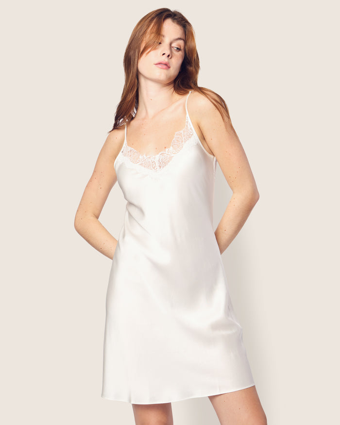 A woman with long red hair wears Petite Plumes Womens Silk Lace Slip Dress in White, featuring a trimmed neckline. Her hands rest in the pockets, capturing minimalist elegance against a plain light-colored background.