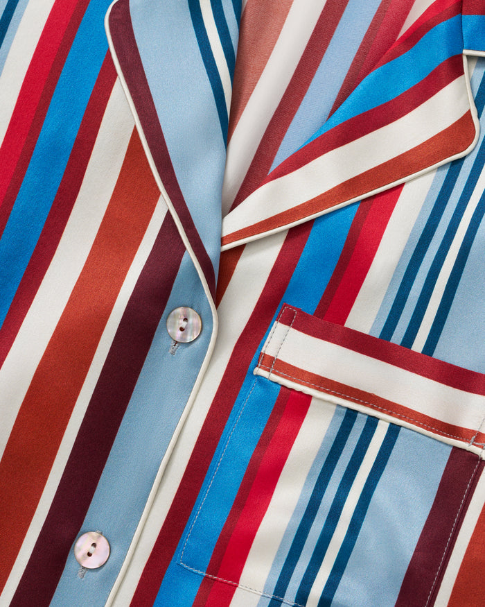 The Women's Silk Pajama Set in Discovery Stripe features long-sleeve, button-up top and matching pants with bold red, blue, brown, and white vertical stripes on smooth silk, displayed on a white background.