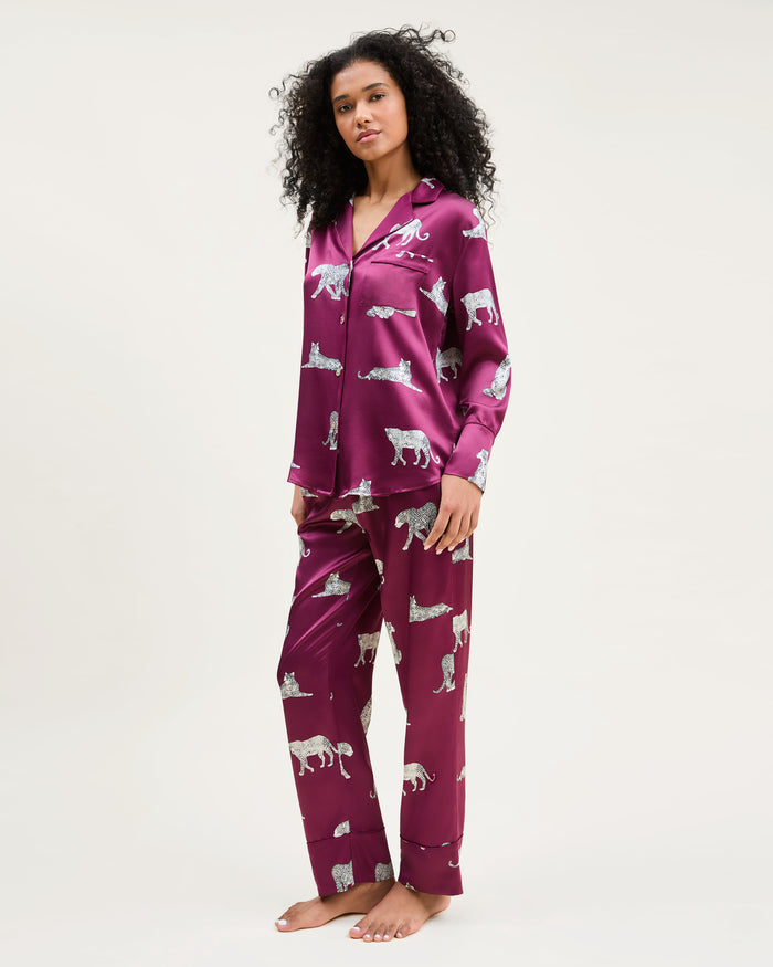 The Women's Silk Pajama Set in Merlot Panthère features a silky, deep magenta button-up shirt and pants adorned with an all-over white illustrated leopard print.