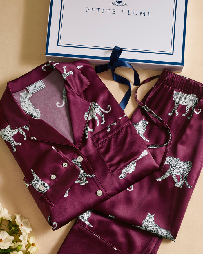 The Women's Silk Pajama Set in Merlot Panthère features a silky, deep magenta button-up shirt and pants adorned with an all-over white illustrated leopard print.