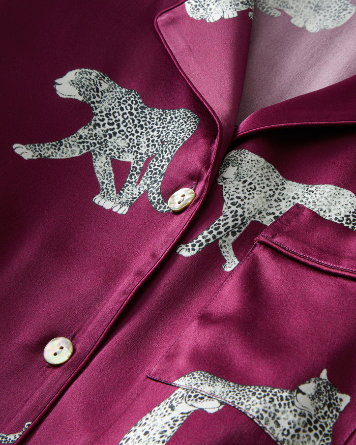 The Women's Silk Pajama Set in Merlot Panthère features a silky, deep magenta button-up shirt and pants adorned with an all-over white illustrated leopard print.