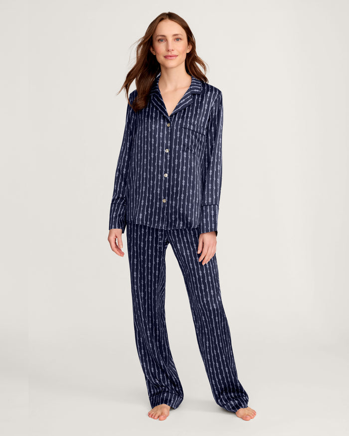 A woman stands barefoot against a plain light background, wearing the Women’s Silk Pajama Set in Nautical Knots—a matching navy blue ensemble featuring a striped pattern, long-sleeve button-up top, and loose-fitting pants.