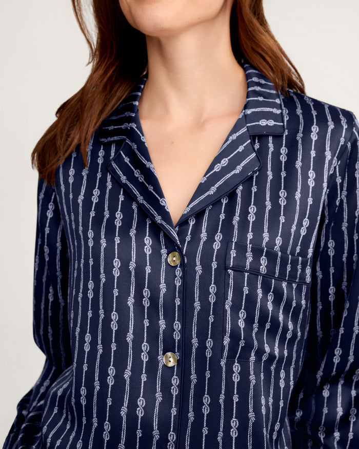 A woman models the Women’s Silk Pajama Set in Nautical Knots, featuring a navy blue, silky top with white vertical stripes and chain patterns, a collar, front pocket, and button closures; her head is tilted and partially out of frame.