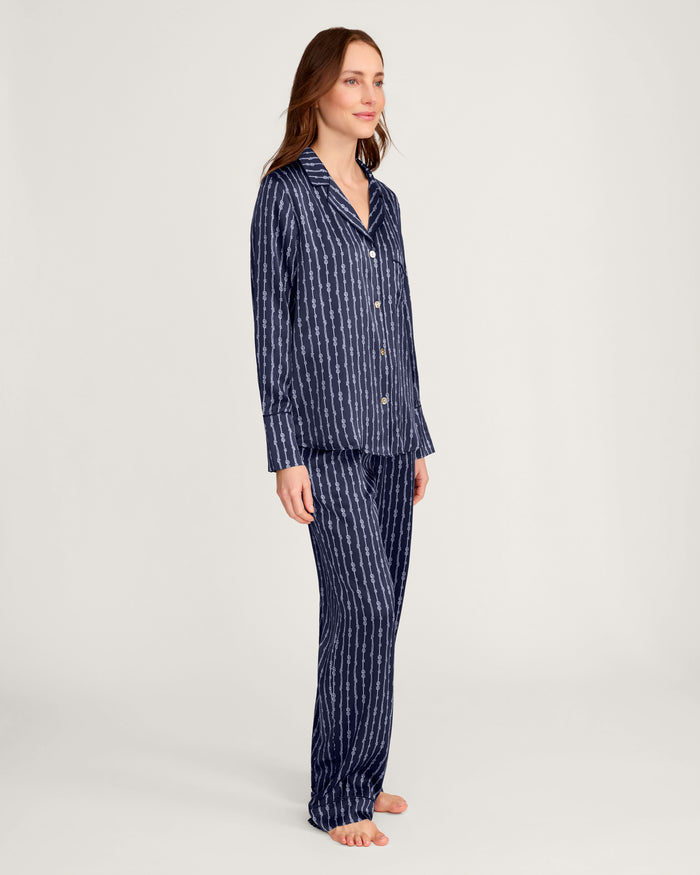 A woman with light skin and long brown hair stands barefoot against a plain background, smiling slightly to the side. She is wearing the Women’s Silk Pajama Set in Nautical Knots, which features matching navy blue long-sleeve top and pants with white vertical stripes.