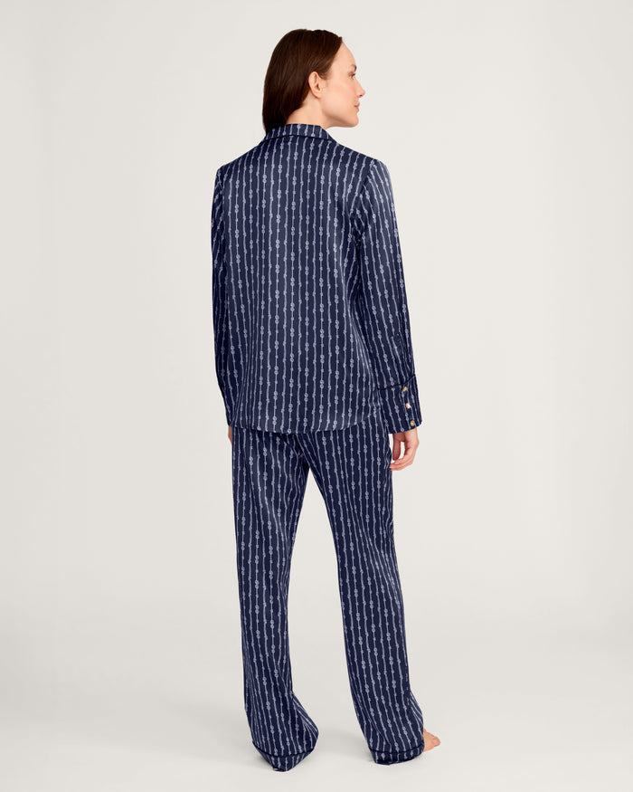 A person stands barefoot, facing away, wearing the Women’s Silk Pajama Set in Nautical Knots—a matching navy blue set featuring a vertical white geometric pattern—against a plain light background.