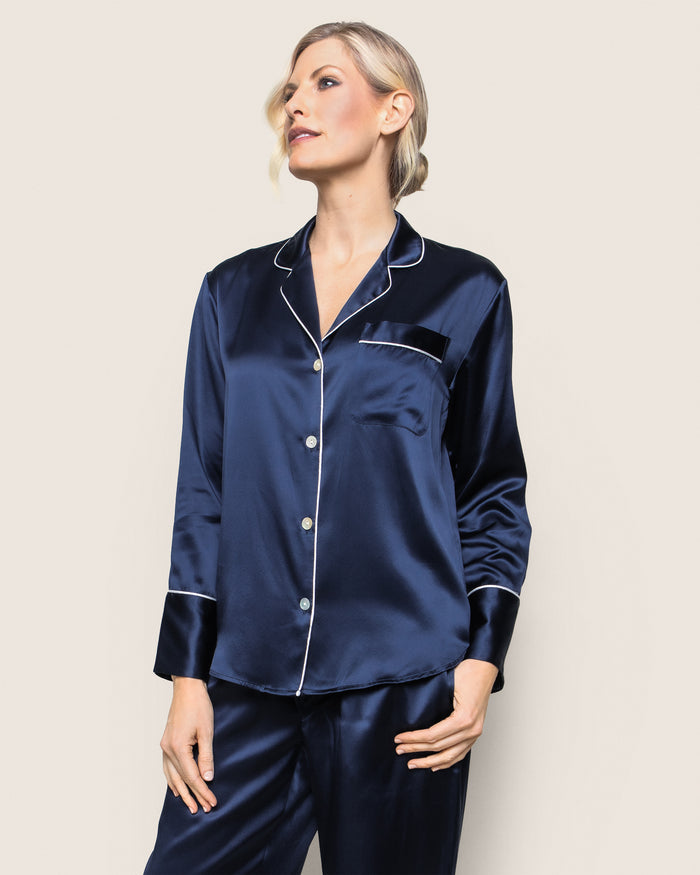 A person with short blonde hair stands against a light background wearing Petite Plumes Womens Silk Pajama Set in Navy. This luxury sleepwear has a timeless silhouette, white piping, and a breast pocket. They appear to be looking to the side.