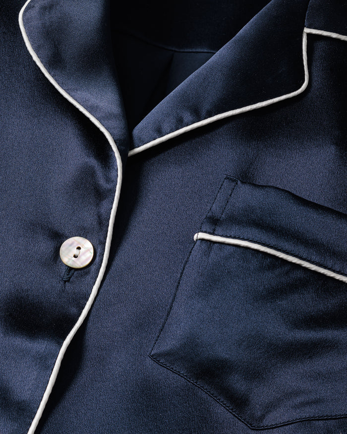 The Womens Silk Pajama Set in Navy by Petite Plume features a close-up of the navy blue silk top with white piping along the collar, pocket, and edges. This luxury sleepwear includes a front pocket and white button, offering a timeless silhouette and luxurious silk texture.