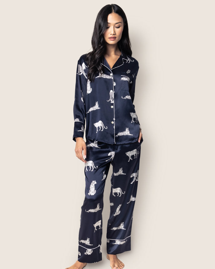 A person with long black hair wears Petite Plumes Womens Silk Pajama Set in Panthère de Nuit, featuring elegant navy blue pajamas adorned with panther prints. Against a plain backdrop, they confidently pose with one hand in the pocket, embodying luxury sleepwear charm.