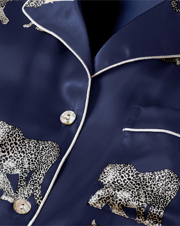 The Womens Silk Pajama Set in Panthère de Nuit by Petite Plume is navy blue with a Mulberry silk button-up shirt and pants. It features white jaguars in various poses and has white piping on the collar, cuffs, and pocket, offering elegant luxury sleepwear.