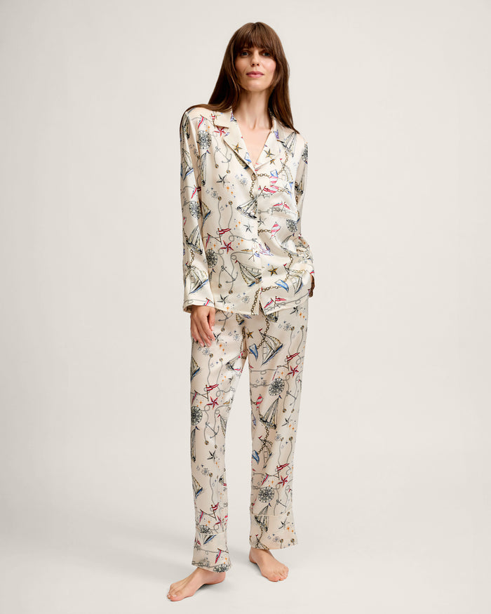 A woman with long brown hair stands barefoot against a plain light background, smiling slightly as she wears the Women’s Silk Pajama Set in Regatta Royale—a cream-colored, long-sleeve pajama set featuring a colorful abstract print.