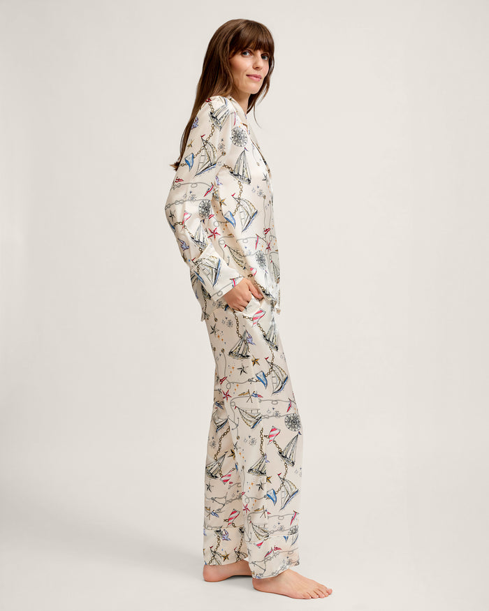 A person stands sideways, smiling, wearing the Women’s Silk Pajama Set in Regatta Royale—a light-colored ensemble featuring a long-sleeve top with a playful print of umbrellas and flowers—against a plain background.