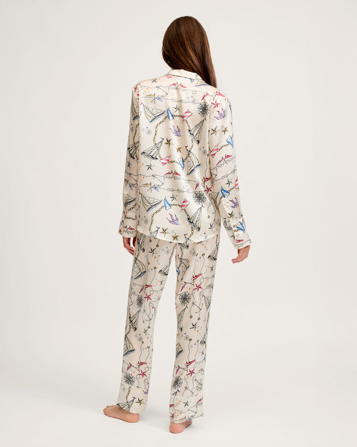 A woman with long brown hair stands barefoot, facing away, wearing the Women’s Silk Pajama Set in Regatta Royale—cream-colored satin pajamas featuring a multicolored star and constellation pattern on both the long-sleeve top and matching pants—against a plain background.