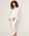 A curly-haired individual, barefoot, stands relaxed and smiling against a plain background in a Petite Plume Womens Silk Pajama Set, white with periwinkle piping. The top features buttons and a chest pocket.