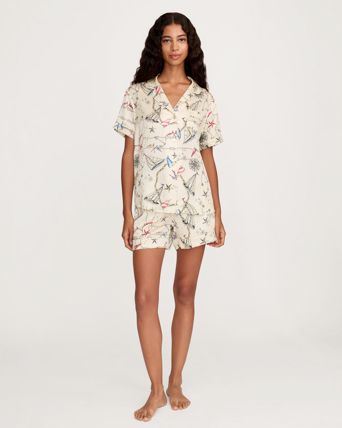 A woman with long curly hair stands barefoot against a plain white background, wearing the Women’s Silk Short Sleeve Short Set in Regatta Royale—a matching cream-colored button-up shirt and shorts adorned with a vibrant kite pattern.