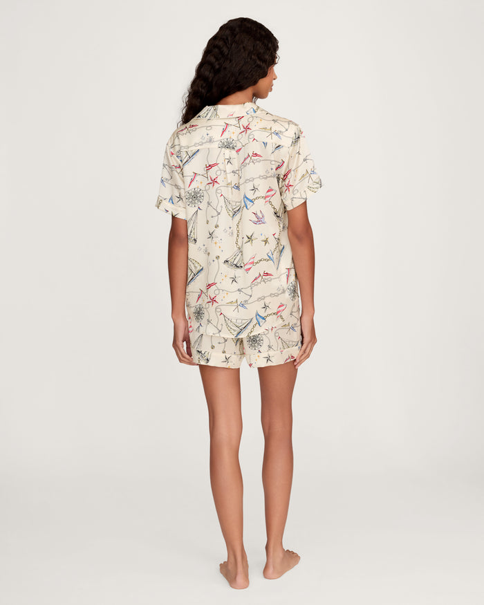 A woman with wavy dark hair stands barefoot, facing away, wearing the Women’s Silk Short Sleeve Short Set in Regatta Royale—a matching pajama set featuring a light background and colorful abstract print. The scene is set in a plain, light-colored studio.