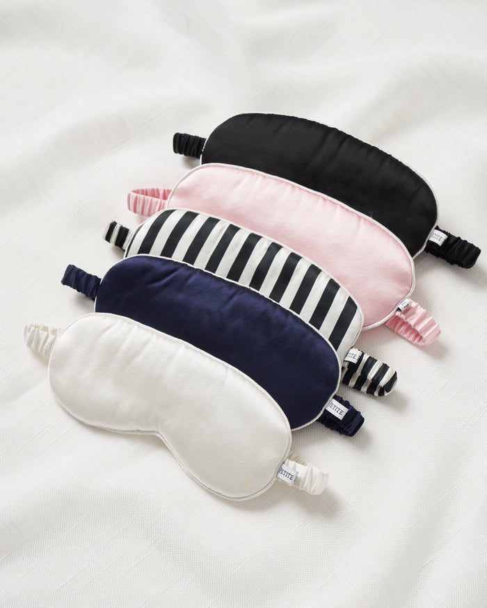 A stack of five premium sleep masks from Petite Plume rests on a white surface, showcasing pure silk textures. From top to bottom: black, pink, black and white stripes, navy blue, and Womens Silk Sleep Mask in White—all featuring an elastic band for secure comfort around the head.